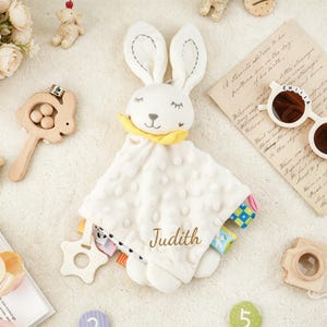 Puede incluir: Una manta de seguridad de felpa blanca con cabeza de conejo, ojos cerrados bordados y pa&ntilde;uelo amarillo. La manta tiene una superficie texturizada con puntos, un mordedor de madera en forma de estrella y el nombre Judith bordado en hilo dorado.