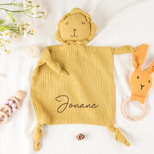 May include: Mustard yellow baby security blanket with a lion head and knotted arms and legs. A matching bunny-shaped teether and a wooden rattle are also present. The blanket is personalized with the name "Jonane".
