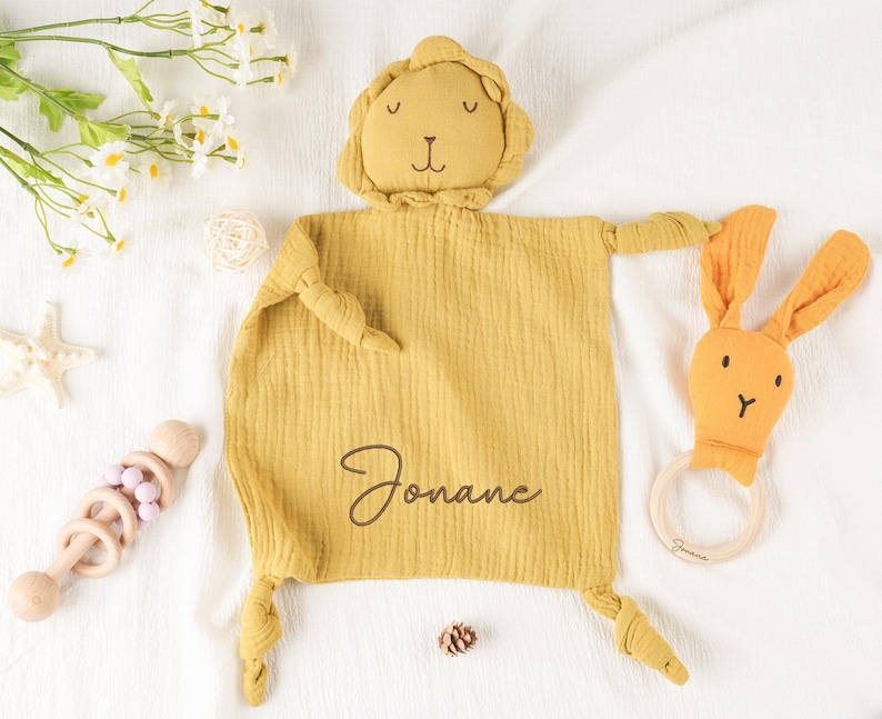 May include: Mustard yellow baby security blanket with a lion head and knotted arms and legs. A matching bunny-shaped teether and a wooden rattle are also present. The blanket is personalized with the name "Jonane".