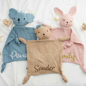 May include: Three baby security blankets with animal heads: a blue koala, a tan bear, and a pink bunny. Each blanket has a name embroidered on it. A wooden rattle and starfish are also visible.