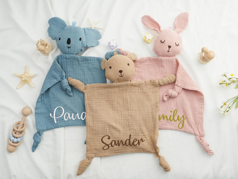May include: Three baby security blankets with animal heads: a blue koala, a tan bear, and a pink bunny. Each blanket has a name embroidered on it. A wooden rattle and starfish are also visible.