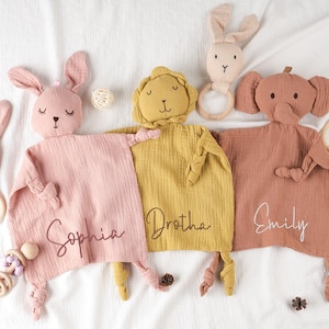 May include: A collection of soft, plush baby comforters in pink, yellow, and brown, each with a different animal head: a bunny, a lion, and an elephant. Each comforter has a name embroidered on it. Additional bunny-shaped teethers are also present.
