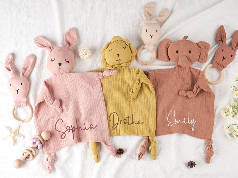 May include: A collection of soft, plush baby comforters in pink, yellow, and brown, each with a different animal head: a bunny, a lion, and an elephant. Each comforter has a name embroidered on it. Additional bunny-shaped teethers are also present.