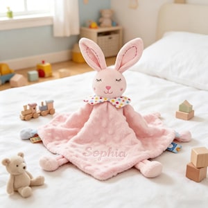 Puede incluir: Una suave manta de seguridad de peluche rosa con una cabeza de conejo, ojos cerrados bordados y una nariz en forma de coraz&oacute;n. La manta tiene una textura con hoyuelos, un cuello de lunares de colores y el nombre Sophia bordado en rosa.