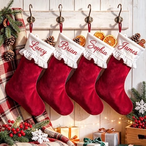 May include: Four red velvet Christmas stockings with white ruffled cuffs, each embroidered with a name: Laecy, Brian, Sophia, and James. The stockings hang from metal hooks on a wooden board, decorated with pine branches and orange slices.