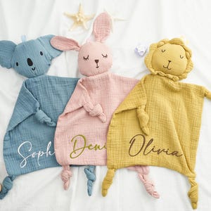 May include: Three baby security blankets in blue, pink, and yellow, each with an animal head and knotted corners. The blue blanket has a koala head, the pink one a bunny, and the yellow one a lion. Each blanket has a name embroidered on it.