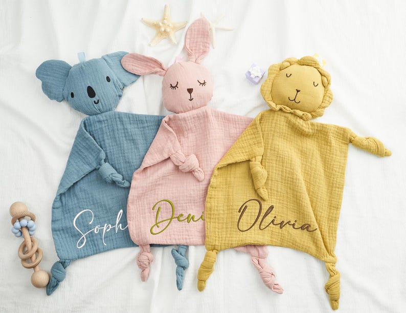 May include: Three baby security blankets in blue, pink, and yellow, each with an animal head and knotted corners. The blue blanket has a koala head, the pink one a bunny, and the yellow one a lion. Each blanket has a name embroidered on it.