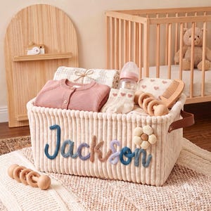 May include: A cream-colored corduroy storage basket featuring the name Jackson in blue, brown, and purple yarn lettering with a fabric flower. The basket contains a pink cardigan, baby socks, a bottle, a wooden comb, and wooden ring toys.