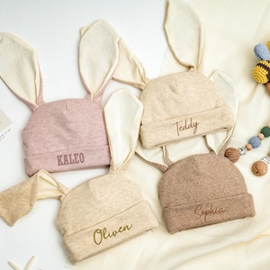 May include: Four beige and pink bunny ear hats with different names embroidered on them: Kaleo, Teddy, Sophia, and Oliven.