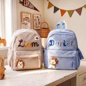 May include: Two corduroy backpacks, one beige and one light blue, featuring vertical stripes. The beige bag displays the name Liam with a small flower, and the blue bag displays the name Daniel. Both have front pockets and plush animal patches.