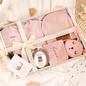 May include: A baby gift set in a white box, featuring pink clothing, a hat, a bunny-shaped toy, and a wooden ornament. The set includes a onesie and a shirt, both with the name "Anna" embroidered. The box is tied with a cream-colored ribbon.