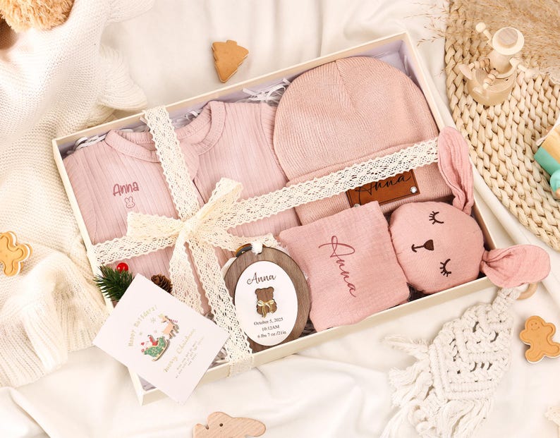 May include: A baby gift set in a white box, featuring pink clothing, a hat, a bunny-shaped toy, and a wooden ornament. The set includes a onesie and a shirt, both with the name "Anna" embroidered. The box is tied with a cream-colored ribbon.
