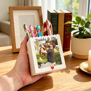 May include: A white square pen holder constructed from building blocks, featuring a printed photograph of a person lifting a child outdoors. The text Daddy & Me is printed above the photo, with a small red heart icon below the image.