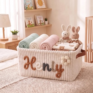 May include: A rectangular cream-colored corduroy storage basket featuring the name Henry in raised, multi-colored yarn lettering and a fabric flower appliqué. The basket contains three rolled blankets in mint, white, and pink, plus two plush rabbit toys.