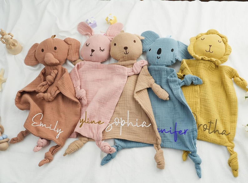 May include: Five soft, plush baby security blankets in the shape of animals: an elephant, a bunny, a bear, a koala, and a lion. Each blanket is a different color, with a name embroidered on the front. The elephant is brown, the bunny is pink, the bear is tan, the koala is blue, and the lion is yellow.