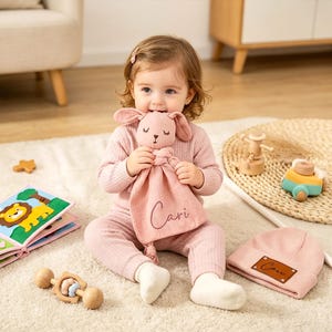 May include: A baby girl in pink clothing holds a pink bunny lovey with the name "Cari" embroidered on it. A matching pink beanie with a brown patch and a wooden rattle are nearby. A colorful soft book and other toys are also visible.