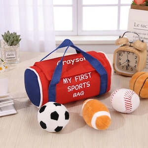 May include: A red and blue sports bag with the text "MY FIRST SPORTS BAG" and "Grayson" in white. The bag has blue handles and is next to a soccer ball, football, baseball, and basketball. A wooden alarm clock and a small plant are also visible.