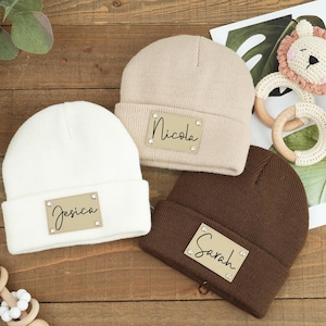Puede incluir: Tres gorros de punto en beige, blanco y marrón con etiquetas de nombre personalizadas. Las etiquetas son de cuero marrón con borde dorado y tienen los nombres "Nicola", "Jessica" y "Sarah" escritos en negro.