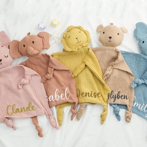 May include: A collection of soft, plush baby comforters in various animal shapes, including a bunny, elephant, lion, bear, and koala. Each comforter is a different pastel color and has a name embroidered on it.