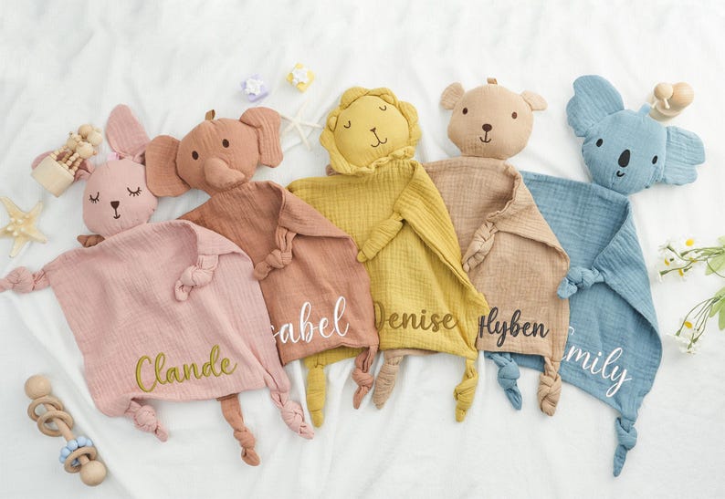 May include: A collection of soft, plush baby comforters in various animal shapes, including a bunny, elephant, lion, bear, and koala. Each comforter is a different pastel color and has a name embroidered on it.