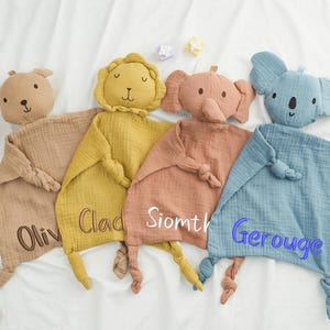 May include: Four soft, animal-shaped baby security blankets in tan, yellow, peach, and blue. Each blanket features an embroidered name and a knotted corner. The animals include a bear, lion, elephant, and koala.