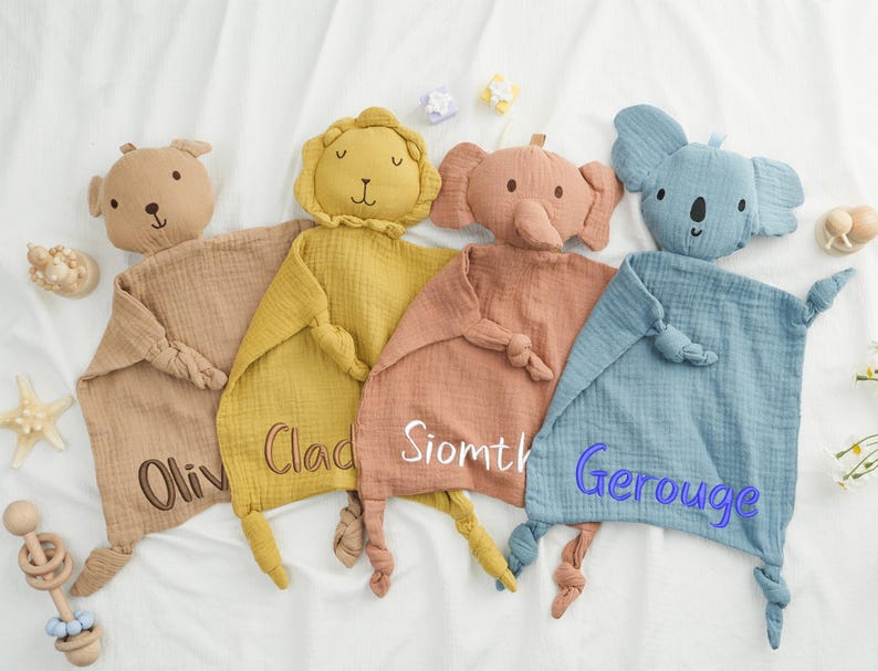 May include: Four soft, animal-shaped baby security blankets in tan, yellow, peach, and blue. Each blanket features an embroidered name and a knotted corner. The animals include a bear, lion, elephant, and koala.
