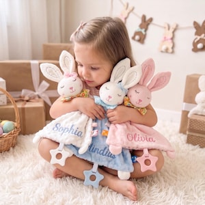 Puede incluir: Tres mantitas de apego de conejo de peluche con los nombres bordados Sophia, Olivia y un tercer nombre. Cada una presenta una cabeza de conejo suave con ojos cerrados, un lazo pequeño y un mordedor de plástico en forma de estrella.