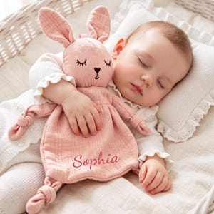 May include: A sleeping baby cuddles a pink bunny comforter in a white wicker bassinet. The bunny has long ears, closed eyes, and the name "Sophia" embroidered on the dress. The baby is wearing a white outfit with ruffled edges.