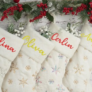 May include: Four white Christmas stockings with snowflake patterns and personalized names: Selina, Olivia, Cerlin, and Anyla. The names are embroidered in red and yellow thread.
