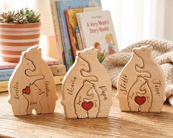 Puede incluir: Tres figuras de rompecabezas de madera clara que representan una familia de osos de pie. Cada rompecabezas de oso tiene nombres grabados en los lados y un pequeño corazón de madera pintado de rojo sostenido por los osos más pequeños.