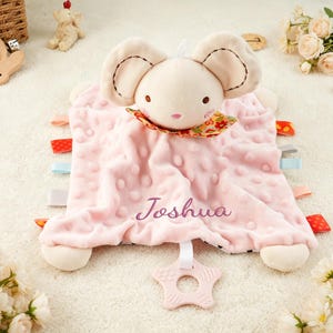 Puede incluir: Una manta de seguridad para beb&eacute; de color rosa suave con una cabeza de rat&oacute;n de peluche, orejas grandes, un cuello floral y el nombre Joshua bordado en morado. La manta tiene una textura de puntos, etiquetas de tela de colores y un mordedor en forma de estrella.