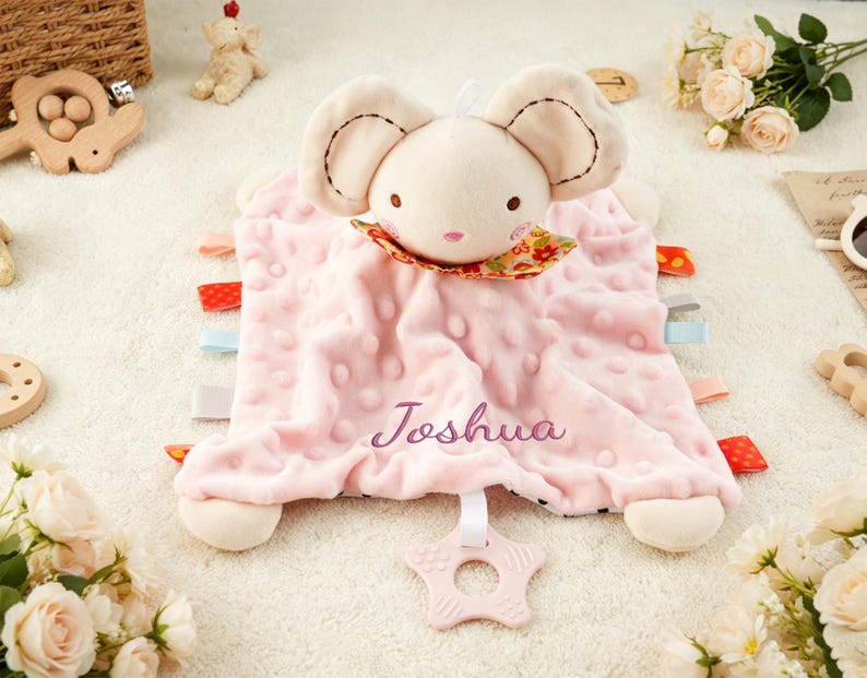 Puede incluir: Una manta de seguridad para beb&eacute; de color rosa suave con una cabeza de rat&oacute;n de peluche, orejas grandes, un cuello floral y el nombre Joshua bordado en morado. La manta tiene una textura de puntos, etiquetas de tela de colores y un mordedor en forma de estrella.
