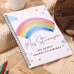 May include: Spiral-bound notebook with a watercolor rainbow design. The cover features the name "Mrs Stevenson" and the text "BIG HEARTS SHAPE LITTLE MINDS". Pastel-colored pencils and a white mug are visible.