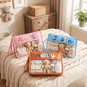May include: Three rectangular zipped pouches in pink, blue, and orange, each featuring a clear front panel and a personalised name in plush lettering. Inside each pouch are baby essentials including a small teddy bear, a bottle, and clothing.