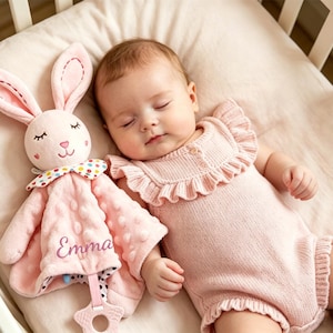 Puede incluir: Una manta de seguridad de peluche rosa con forma de conejo, tejido texturizado con relieve, un lazo de cuello con lunares de colores y el nombre Emma bordado en cursiva. Incluye un mordedor de silicona con forma de estrella en la base.