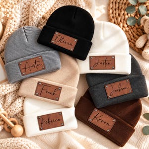 Könnte beinhalten: Eine Sammlung von sieben Strickmützen in Schwarz, Grau, Weiß, Beige und Braun. Jede Mütze hat ein rechteckiges braunes Kunstleder-Etikett, das mit vier Metallnieten befestigt ist. Die Etiketten sind mit den Namen Austin, Oliven, Justin, Frank, Jackson, Rebeica und Keven in schwarzer Schreibschrift personalisiert.