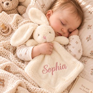 Op de afbeelding: Een crèmekleurig pluche knuffeldoekje met een knuffelkonijnenkop met lange oren en zachte stof. Het doekje is geborduurd met de naam Sophia in roze schrijfletters. Het item is gemaakt van een pluizig, gestructureerd materiaal.