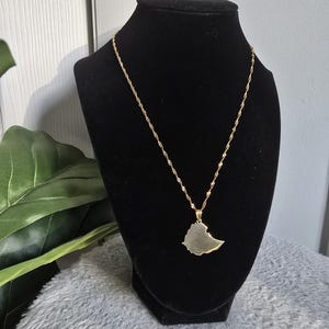 Puede incluir: Un collar de cadena dorada con un colgante con forma del contorno de Etiopía. El collar se muestra en un soporte de terciopelo negro.