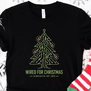 May include: Black t-shirt with a neon green Christmas tree design made of circuit board lines. The text "WIRED FOR CHRISTMAS - CIRCUITS OF JOY" is below the tree. Red and white striped socks with a Santa Claus face are next to the shirt.