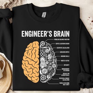 Könnte beinhalten: Schwarzes Sweatshirt mit dem Text "ENGINEER'S BRAIN" und einer Grafik eines Gehirns, das in zwei Hälften geteilt ist. Eine Hälfte ist hellorange, die andere ein detailliertes mechanisches Design in Weiß. Enthält Beschriftungen wie "Problem Solving Function" und "Coffee Location Sensor".