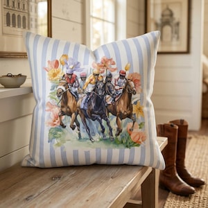 May include: Square throw pillow with a watercolor print of a horse race scene. The pillow has a light blue and white striped background. The artwork features jockeys in colorful silks riding brown and black horses, surrounded by flowers.