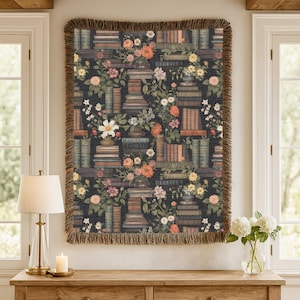 May include: A rectangular woven throw blanket featuring a dark background with a repeating pattern of stacked books and various colourful floral arrangements. The blanket has a textured surface and is finished with a fringed edge on all sides.