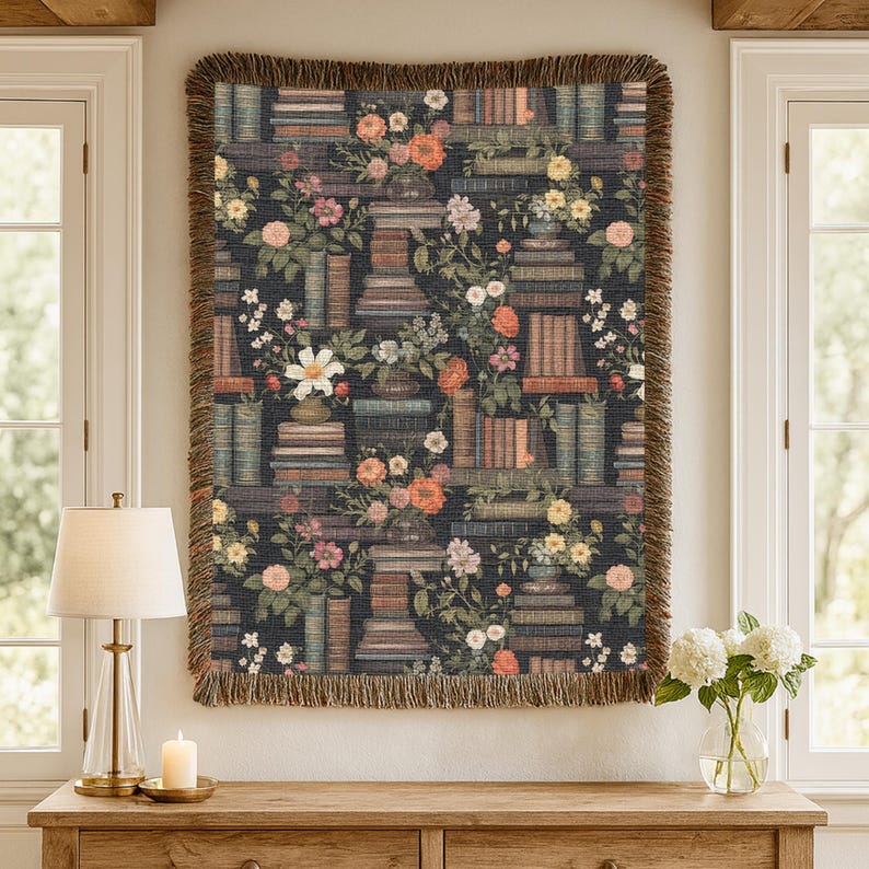 May include: A rectangular woven throw blanket featuring a dark background with a repeating pattern of stacked books and various colourful floral arrangements. The blanket has a textured surface and is finished with a fringed edge on all sides.
