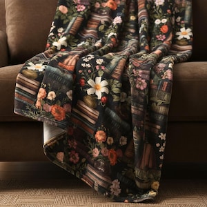 May include: A black throw blanket featuring a repeating pattern of stacked vintage books interspersed with colourful floral arrangements including white daisies, orange poppies, and pink blossoms, creating a dense botanical and literary design.