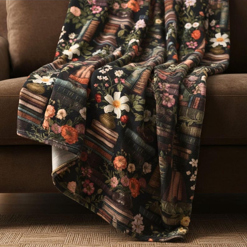 May include: A black throw blanket featuring a repeating pattern of stacked vintage books interspersed with colourful floral arrangements including white daisies, orange poppies, and pink blossoms, creating a dense botanical and literary design.