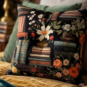 May include: Cushion with a black background featuring a floral and book design. The cushion has stacks of books, vases, and various flowers in shades of orange, white, and green. The cushion is on a gold-coloured surface.
