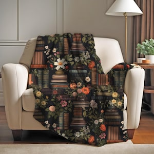 May include: A soft throw blanket featuring a dark black background with a repeating pattern of stacked vintage books and colourful floral arrangements including white daisies, orange roses, and small yellow blossoms interspersed throughout.