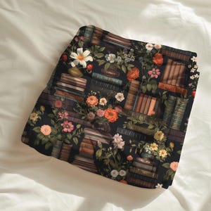 Peut inclure: Tissu sombre avec un motif de livres et de fleurs. Le motif pr&eacute;sente des piles de livres, des vases et des fleurs aux teintes orange, rose et blanc. Le tissu est pos&eacute; sur un fond blanc.