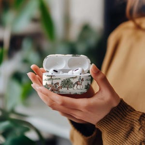 May include: White AirPod case with a toile pattern featuring riders on horseback, trees, and foliage. The case is open, revealing white earbuds. The case is held in a person's hands.