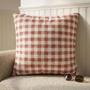 May include: A square decorative pillow with a red and white gingham pattern. The pillow is resting on a beige upholstered surface, with a pair of sunglasses nearby. The pillow has a soft, inviting appearance.